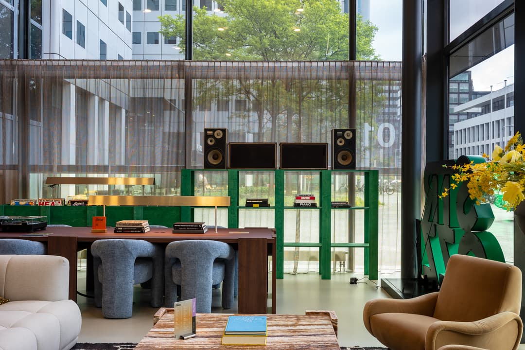 Modern lounge with plush seating, a wooden table, books, and speakers. Large windows reveal a view of urban greenery outside.