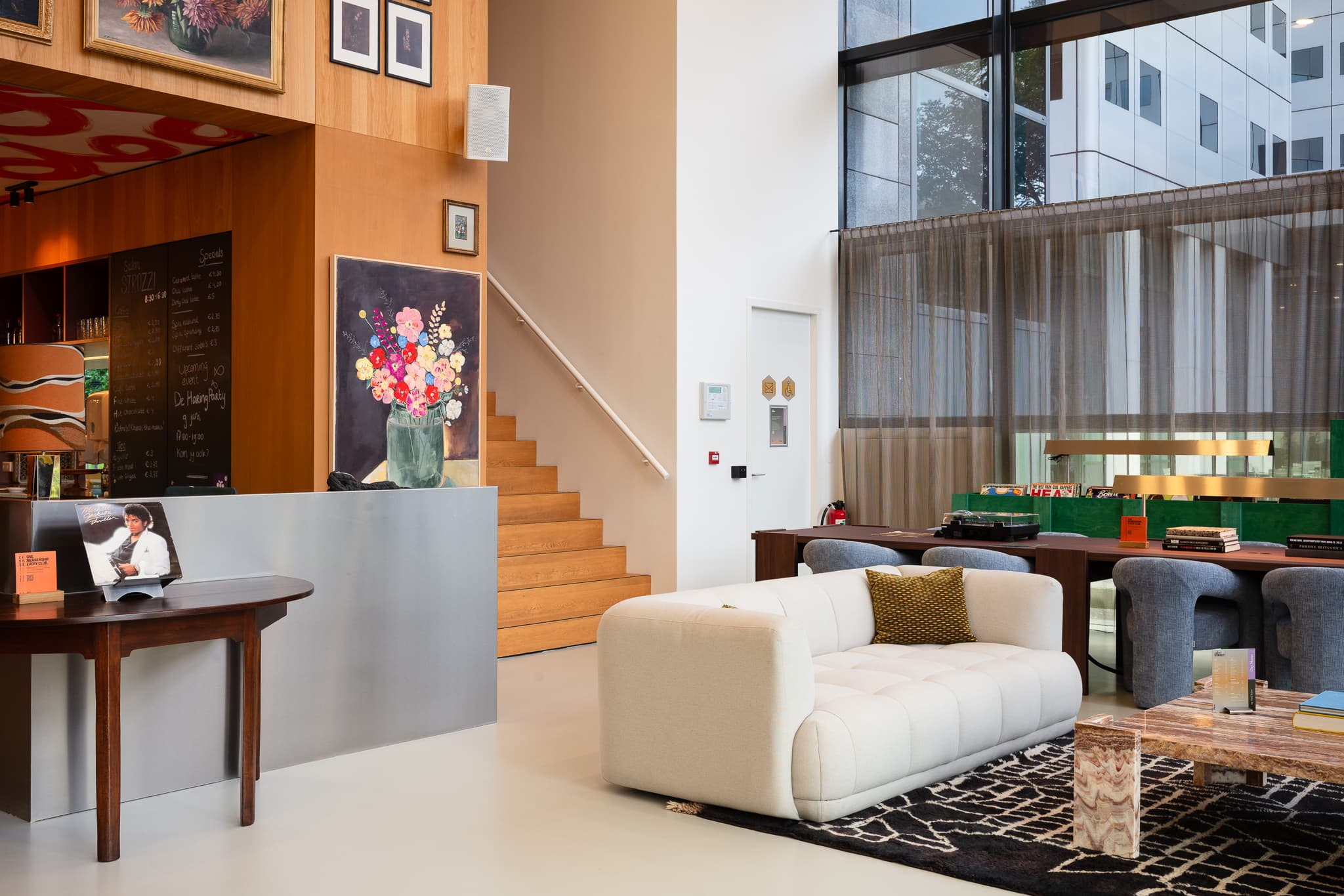 Modern lobby with a white sofa, patterned rug, artwork, wooden stairs, and a desk with books and decor. Natural light from large windows.