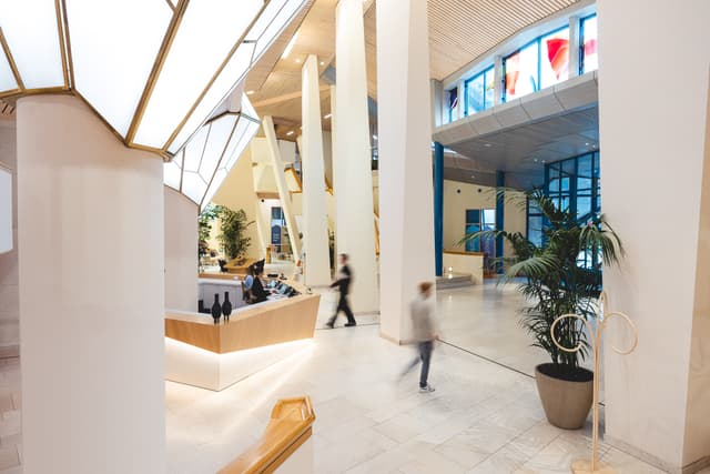 Modern lobby with high ceilings, large windows, and geometric light fixtures. People walk by a reception desk. Indoor plants enhance the space.