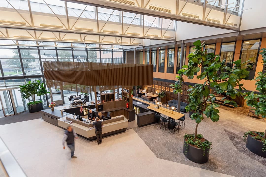 Spacious modern atrium with a central café, large windows, indoor plants, and people walking and sitting at tables.