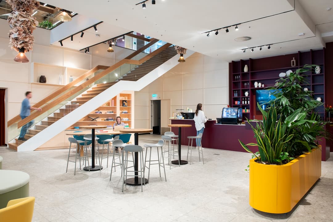 Modern office lounge with wooden staircase, barista counter, and seating area. Potted plants and shelves with books and decor enhance the space.