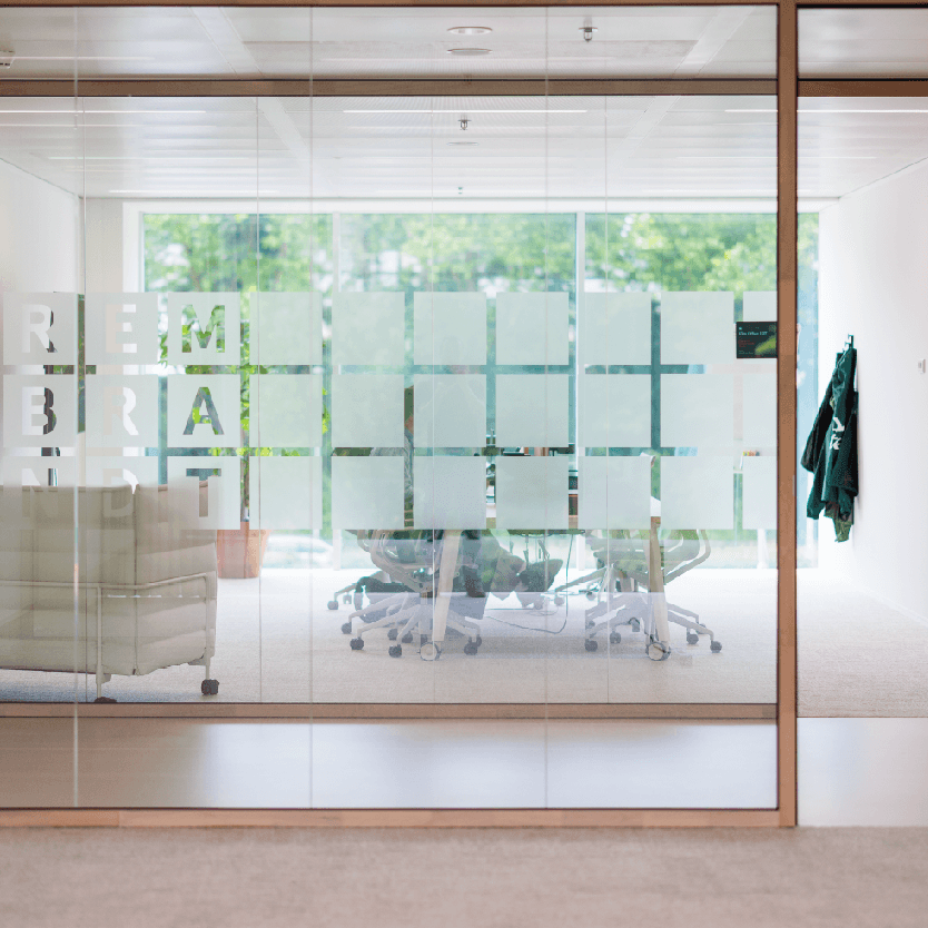 Glass-walled office with frosted squares, chairs around a table, a potted plant, and jackets hanging. Greenery visible outside.
